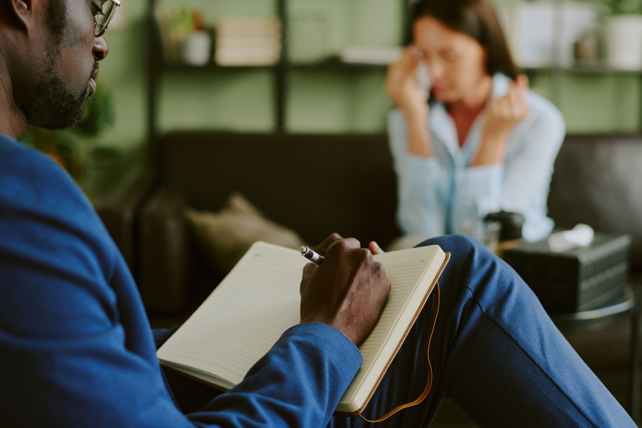 Therapist writing notes during counseling session with client who gestures emotionally in office setting. Shelving unit with decor visible in background
